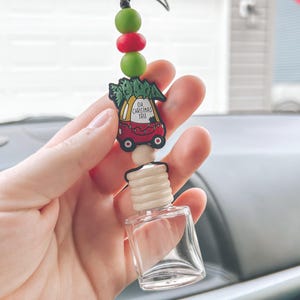 May include: A car air freshener with a clear glass bottle and a wooden cap. The charm features a red car carrying a Christmas tree, with the text "Oh, Christmas Tree." The charm is strung with green, red, and beige beads.