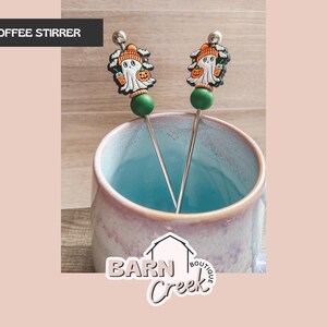 May include: Two Halloween-themed coffee stirrers with a ghost design. Each stirrer features a silver stem, a green bead, and a ghost wearing an orange hat and holding a pumpkin. The stirrers are in a blue and purple mug.
