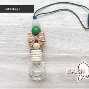 May include: An air freshener diffuser with a clear glass bottle and a wooden top. The diffuser is decorated with a football-shaped bow, green and white beads, and a black cord. The word "DIFFUSER" is in a black rectangle.