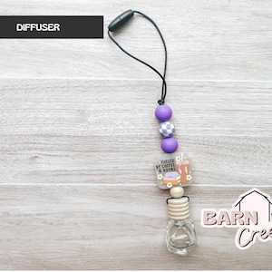 May include: A car air freshener with a black cord, purple beads, and a charm that reads "Fueled by Coffee & Books." The bottom features a clear glass bottle with a wooden top. The word "DIFFUSER" is in a black rectangle.