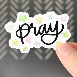 May include: A white sticker with the word "Pray" in black script, surrounded by small pastel-colored flowers and hearts. The sticker is held up against a gray background.