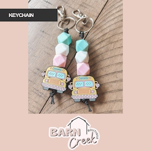 May include: Two keychains with a school bus charm. Each keychain has a series of pastel-colored beads and a silver clasp. The school bus charms are yellow with blue and white details, and the word "SCHOOL" is visible.