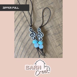 May include: Two black zipper pulls with a white treble clef design and blue beads. The text "ZIPPER PULL" is in a black rectangle at the top. The Barn Creek Boutique logo is at the bottom.