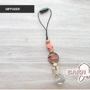May include: A car air freshener with a black cord and a decorative design. The design includes wooden beads, a pink geometric bead, and a round charm with the text "Wake up and smell the coffee". A small glass bottle is at the bottom.