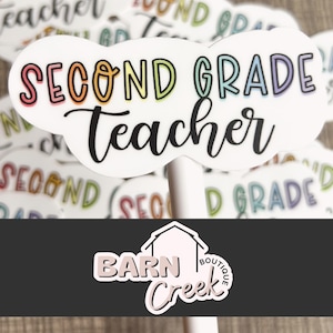 May include: A white cloud-shaped sticker with the text "SECOND GRADE Teacher" in rainbow colors. The sticker is part of a set of stickers with various teacher-related designs. The sticker is from Barn Creek Boutique.
