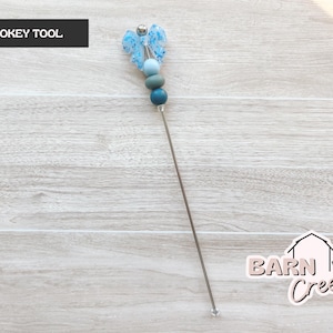 May include: A pokey tool with a butterfly design and blue beads. The tool has a long, thin, silver metal shaft. The top of the tool features a butterfly-shaped embellishment with light blue accents and a series of blue beads in varying shades.