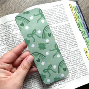 May include: Green bookmark with a pattern of rainbows, shamrocks, hearts, and bows. The bookmark is rectangular and is held over an open book.