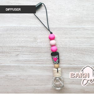 May include: A car air freshener with a black cord and pink, light pink, and wooden beads. The charm features a black and white coffee cup design with a pink heart. The glass bottle is clear and has a wooden top. The word "DIFFUSER" is in a black rectangle.