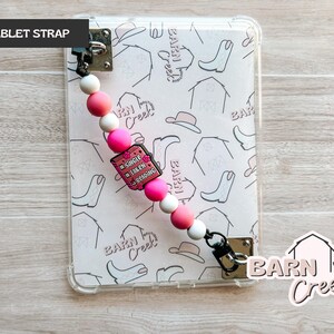 May include: A tablet strap with a pink, white, and black beaded design. The strap features a charm that reads "Single, Taken, Reading." The tablet case has a pattern of cowboy boots, hats, and barns. The words "Barn Creek Boutique" are also visible.