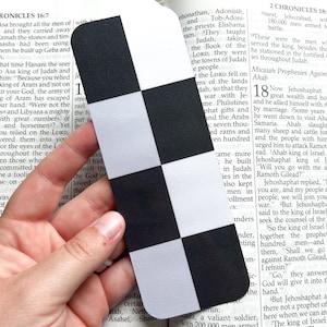 May include: A black and white checkered bookmark is held over an open book. The bookmark has a rounded top and is made of paper. The book is open to a passage from the Bible.