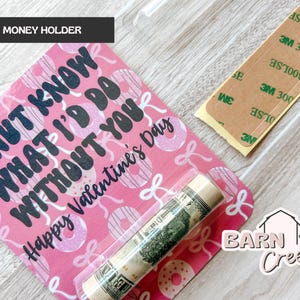 May include: A pink money holder with a Valentine's Day theme. The holder features a donut pattern and the text "Donut Know What I'd Do Without You". A rolled-up bill is visible inside a clear plastic sleeve.