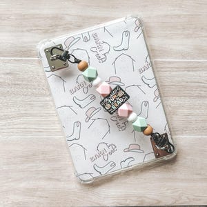 May include: A clear phone case with a white patterned design featuring cowboy boots, hats, and the text "Barn Creek." A beaded lanyard in pastel colors with the text "Fun to Read" is attached to the case.