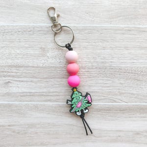 May include: A Christmas-themed keychain with a silver clasp and ring. The keychain features three pink beads and a green Christmas tree charm holding a pink drink. Black cord tassels hang below the charm.