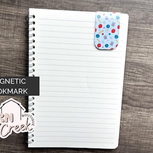May include: A magnetic bookmark with a school-themed pattern of red apples, blue pencils, and other school supplies on a light blue background. The bookmark is attached to a lined notebook. The words "MAGNETIC BOOKMARK" are in a black rectangle.