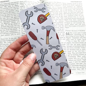 May include: A gray bookmark with a repeating pattern of tools, including wrenches, hammers, screwdrivers, and tape measures. The tape measures have "#1 DAD" printed on them. The bookmark is held over an open book.