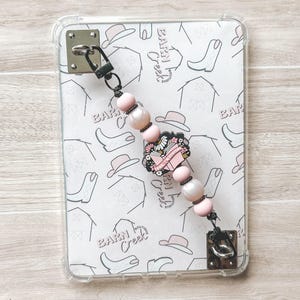 May include: Clear phone case with a white pattern of hats, boots, and barns. A beaded charm with pink beads, pearls, and a floral design is attached. The charm has a black clasp and a metal square attachment.