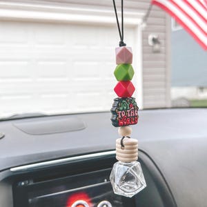 May include: A car air freshener hanging from a black string. It features a series of colorful beads in pink, green, and red, followed by a charm that reads "Joy to the World." Below is a wooden bead and a small glass bottle.