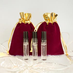 May include: Three clear glass perfume vials with black caps are in front of two claret velvet drawstring bags with gold satin ribbon closures. The vials have the text "Crystal 23" printed on them. The background is white.