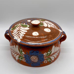 May include: A round, lidded ceramic pot with a reddish-brown base and lid. The lid and base are decorated with white, blue, and green floral patterns. The pot has two small handles and a small knob on the lid.