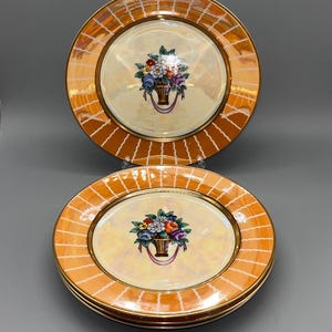 May include: Set of decorative plates with a floral basket design. The plates have a cream-colored center with a gold rim and an orange, grid-patterned border. The floral basket features colorful flowers. The set includes four plates.