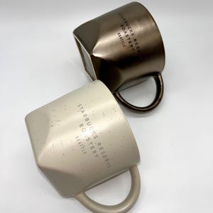 May include: Two Starbucks Reserve Roastery mugs. One is a metallic bronze color, the other is a speckled beige. Both mugs have a geometric design and a curved handle. The text "STARBUCKS RESERVE ROASTERY SEATTLE" is printed on the side of each mug.