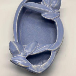 May include: A light blue ceramic decorative tray with a floral design. The tray has an oval shape with a raised rim and a central depression. The floral design is on the sides of the tray. The tray is approximately 10 inches long and 6 inches wide.
