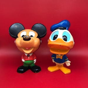 May include: Two vintage Disney character figurines. Mickey Mouse is wearing a red shirt, green pants, and black shoes. Donald Duck is wearing a blue sailor shirt and hat, with orange feet and beak. Both figures have large heads and are set against a red background.