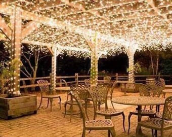 May include: A patio with a wooden pergola covered in white string lights. The patio is furnished with wicker chairs and tables.