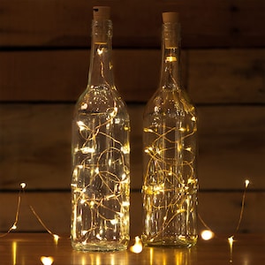 May include: Two clear glass bottles with cork stoppers filled with warm white fairy lights. The lights are strung throughout the bottles, creating a warm and inviting glow.