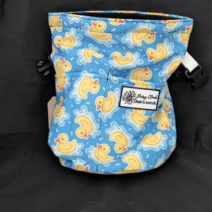 以下が含まれることがあります： 水色のチョークバッグ。黄色いアヒルのラバーダックが水面に浮かぶ模様。バッグは黒い開口部、白いストラップ、そして「Daisy Climb Made in Australia」の文字が入った小さなポケットが付いています。