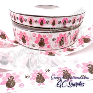 May include: A white grosgrain ribbon spool featuring a repeating pattern of brown turkeys with pink glittery tail feathers and small black swirl accents. The ribbon has a textured ribbed surface and is displayed as a stacked roll with loose ends.