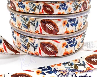 May include: A roll of white ribbon with a pattern of brown footballs, orange flowers, and blue leaves. The ribbon is on a white background.