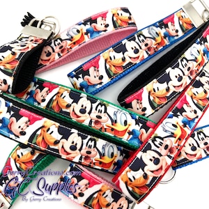 May include: A set of colorful keychains featuring a repeating pattern of Mickey Mouse, Donald Duck, Goofy, and Pluto. The keychains are made of fabric with a black background and white outlines. The keychains are available in pink, blue, green, red, and black.