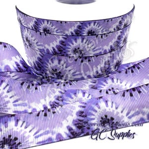 May include: A spool of purple and white tie-dye patterned grosgrain ribbon featuring a repeating sunburst or floral design. The ribbon has a distinct ribbed texture and is wound into a cylindrical roll with a portion extending outward.