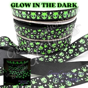 May include: Black ribbon with a repeating pattern of green alien faces, hands, and stars. The text "GLOW IN THE DARK" is at the top. The ribbon is stacked in rolls, with a sample of the glowing ribbon in the dark.