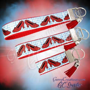 May include: Three keychains with red tassels and silver hardware. The straps feature a red and white gingham pattern with red glittery shoe illustrations. The background has a red, white, and blue gradient.