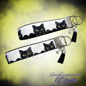 May include: Two black cat keychains with black tassels. The keychains are white with black cat faces peeking out from behind the white fabric. The keychains have silver key rings.