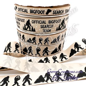 May include: Tan ribbon spools with black Bigfoot silhouettes and text that reads "OFFICIAL BIGFOOT SEARCH TEAM." The ribbon features Bigfoot footprints and forest graphics. The ribbon is suitable for crafting and gift wrapping.