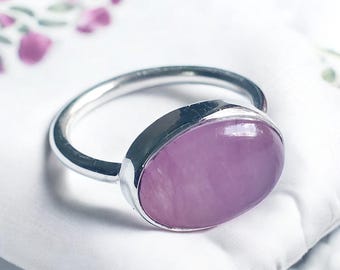 May include: A silver ring with a large oval-shaped pink gemstone. The ring is set on a white fabric background.