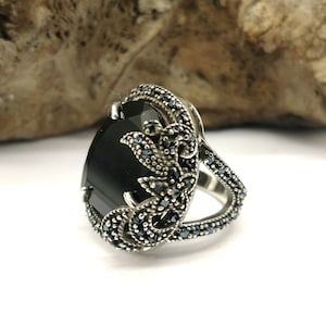 May include: A silver ring with a large black gemstone surrounded by smaller black gemstones. The ring has a floral design.