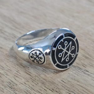 Pode incluir: Um anel de prata com um design de esmalte preto que apresenta uma estrela de oito pontas cercada por um círculo. O anel tem um segundo design menor no lado.