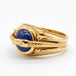 May include: A gold ring with a large, round, deep blue stone. The ring has a textured band and decorative elements, including a snake-like design around the stone. The ring is set against a white background.