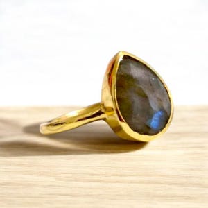 May include: A gold ring with a teardrop-shaped labradorite gemstone. The gemstone has a dark gray color with blue iridescent flashes.