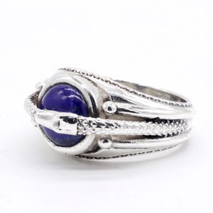 May include: A silver ring featuring a large, oval, deep blue stone. The ring has a detailed, textured design with a snake-like band wrapping around the stone. Small, round accents are placed around the stone setting, adding to the ring's intricate appearance.