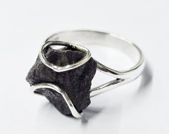 Puede incluir: Un anillo de plata con una piedra de meteorito gris oscuro engastada en el centro. La piedra se mantiene en su lugar por una banda de plata que la envuelve.
