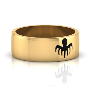 Peut inclure: Une bague dorée à large bande. La bague présente un motif de poulpe noir sur le devant. La bague est présentée sur une surface blanche, avec un reflet de la bague visible en dessous.