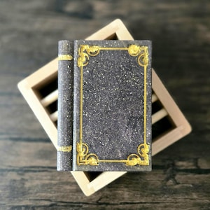 May include: A rectangular, dark gray, speckled object resembling a book with a gold-colored decorative border and spine accents. The object is positioned on top of a light-colored, square, wooden frame with horizontal slats.