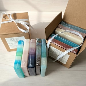 May include: A collection of handcrafted soaps in various colors, including blue, pink, and purple, are displayed. Some soaps are individually wrapped, while others are in a cardboard box tied with a white ribbon. The box has a label that reads "Pretty Soap Co."