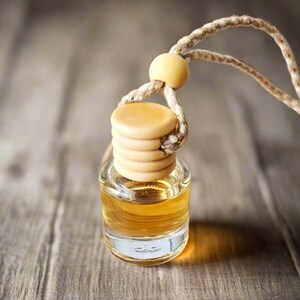 May include: A small, clear glass bottle filled with a golden-colored liquid, topped with a ribbed, light-colored wooden cap. A thick, braided beige cord with a small wooden bead is attached to the cap for hanging this fragrance diffuser.