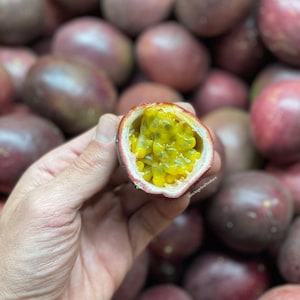 May include: A close-up of a halved passion fruit, revealing its yellow seeds and pulpy flesh. The fruit is held in a hand, surrounded by a pile of whole passion fruits.